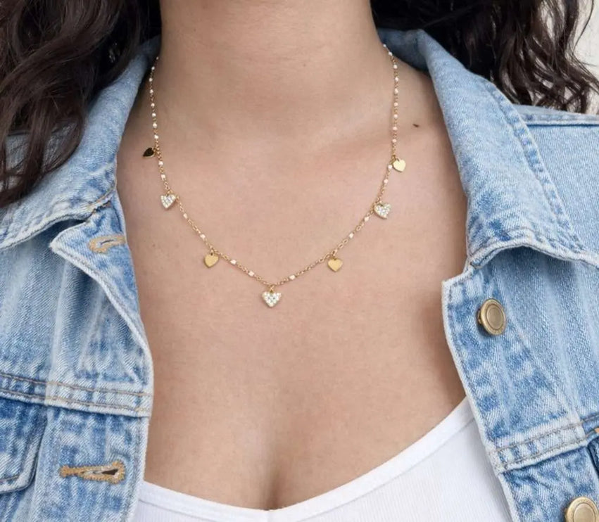 Person wearing a gold necklace with heart-shaped pendants, wearing a denim jacket.